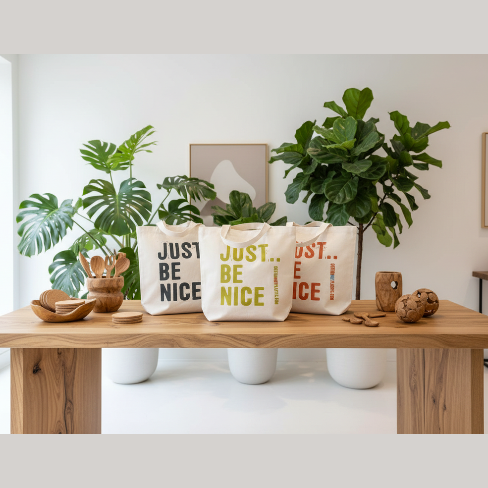 Three tote bags with 'JUST BE NICE' text on a wooden table with plants and decor.
