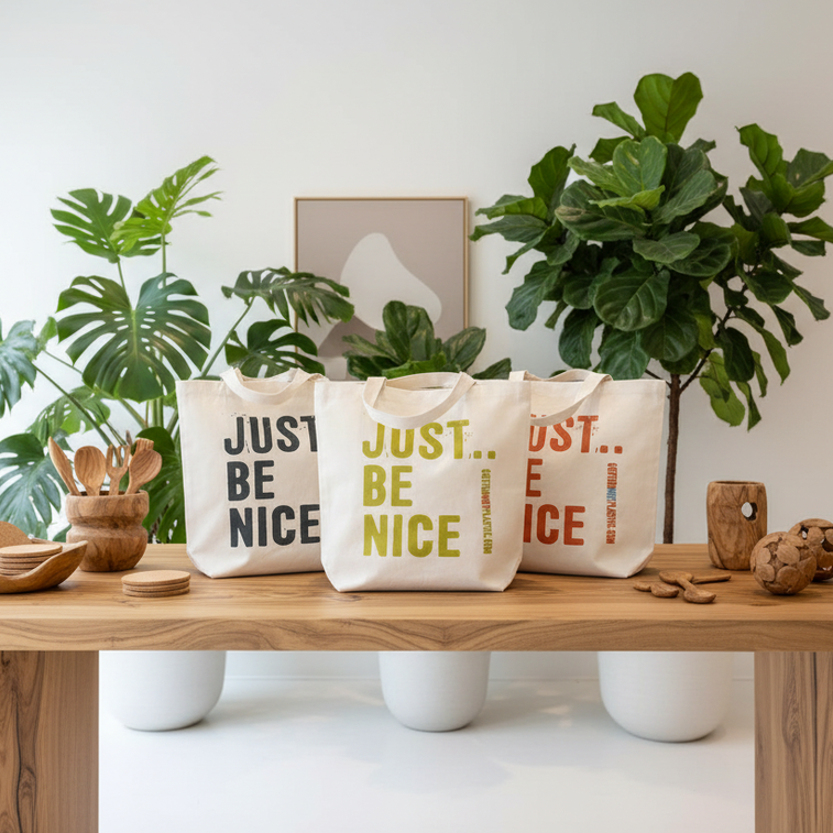 Three tote bags with 'JUST BE NICE' text on a wooden table with plants and decor.