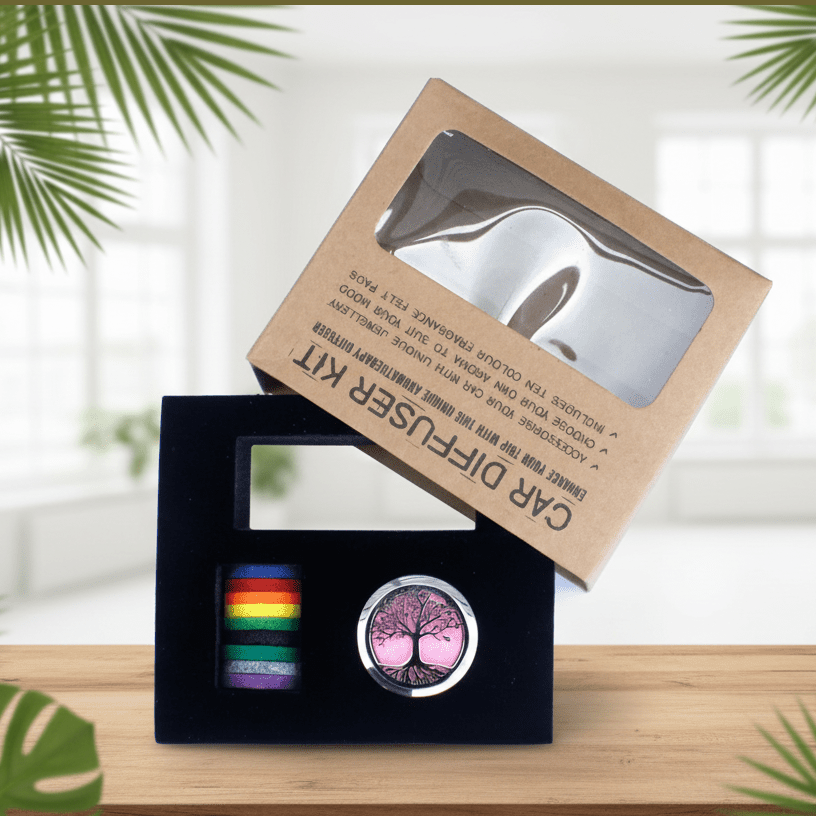 Car diffuser kit in a kraft paper box with a clear window, shown open to reveal a black velvet tray holding a cylindrical rainbow-striped diffuser and a round silver tin with a pink tree design. The main box is propped above the tray on a wooden surface with a blurred indoor background and palm leaves.