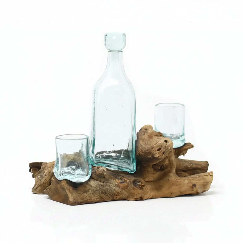 A clear light blue glass bottle with a stopper and two matching small glasses arranged on a natural driftwood base against a white background.