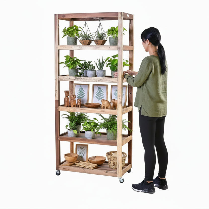 A woman in a green top and black leggings stands beside a tall, rustic wooden rolling shelf with five tiers and caster wheels, styled with eco-friendly home decor and plants against a plain white background.