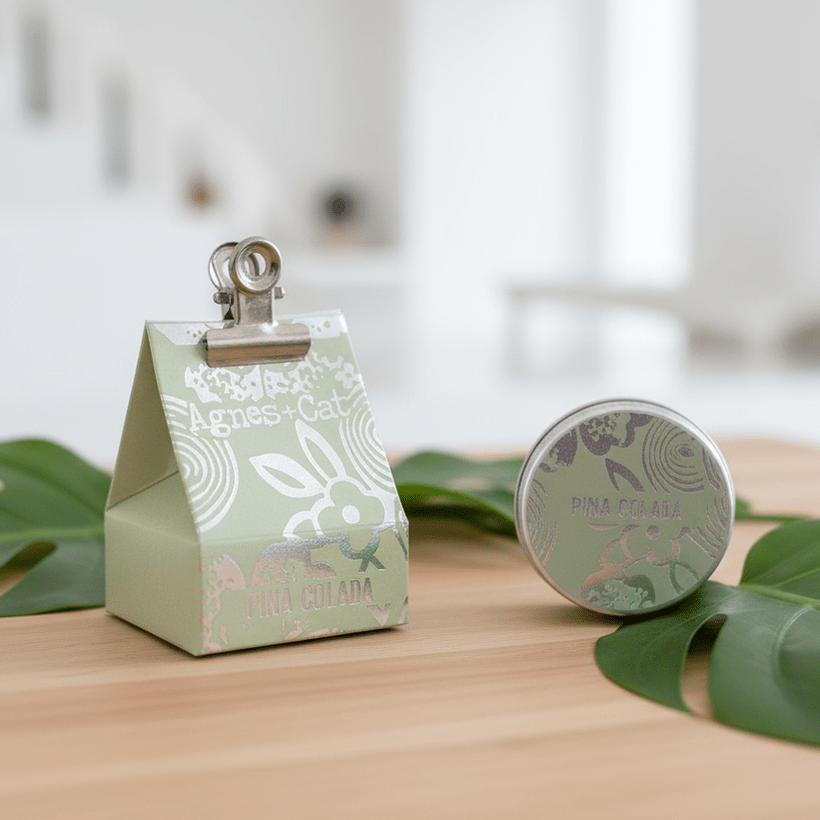 Small green gift box with a clip on top, next to a natural agnes and cat lip balm, on a wooden surface with leaves.