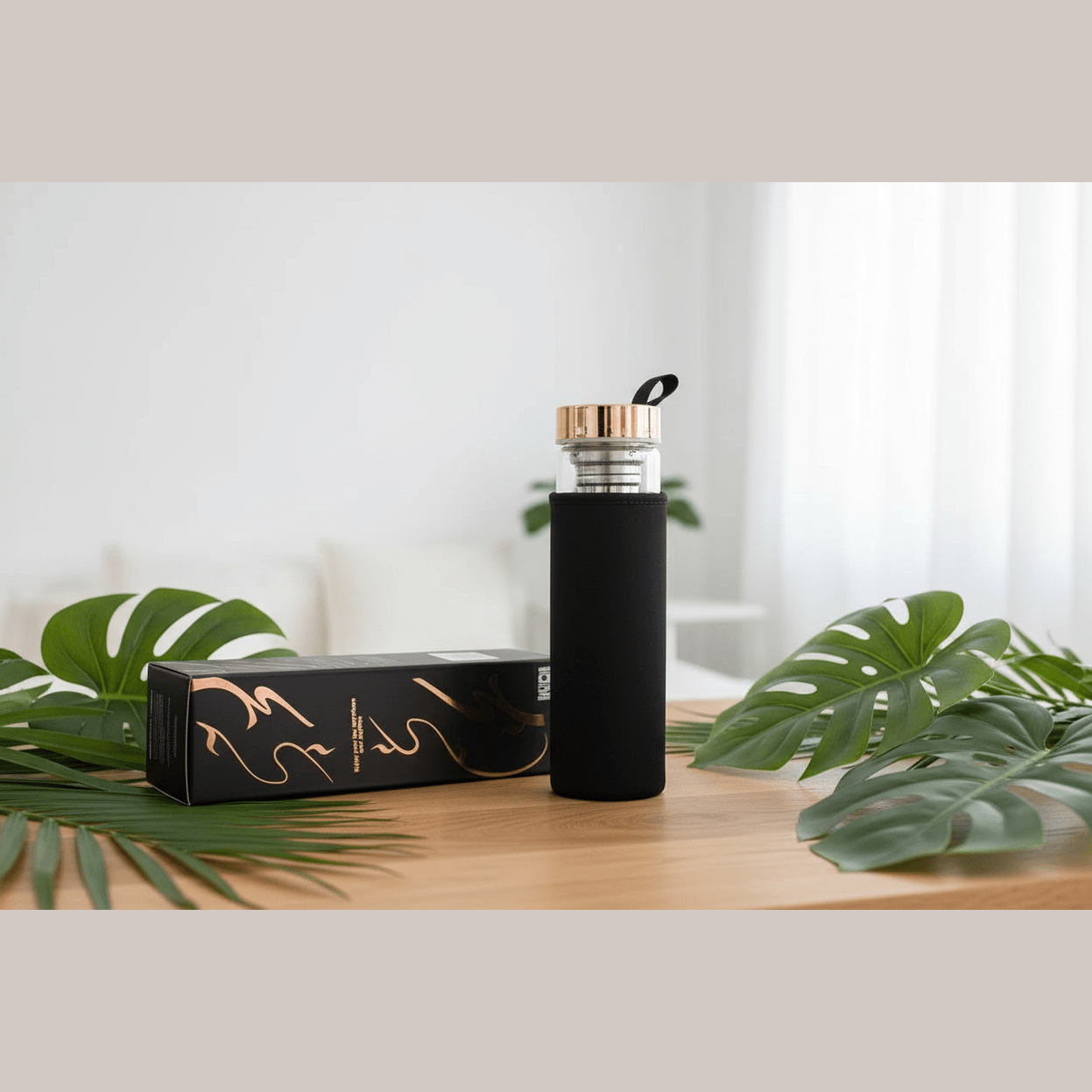 Black insulated bottle with rose gold cap on a wooden surface with green leaves and packaging in the background.