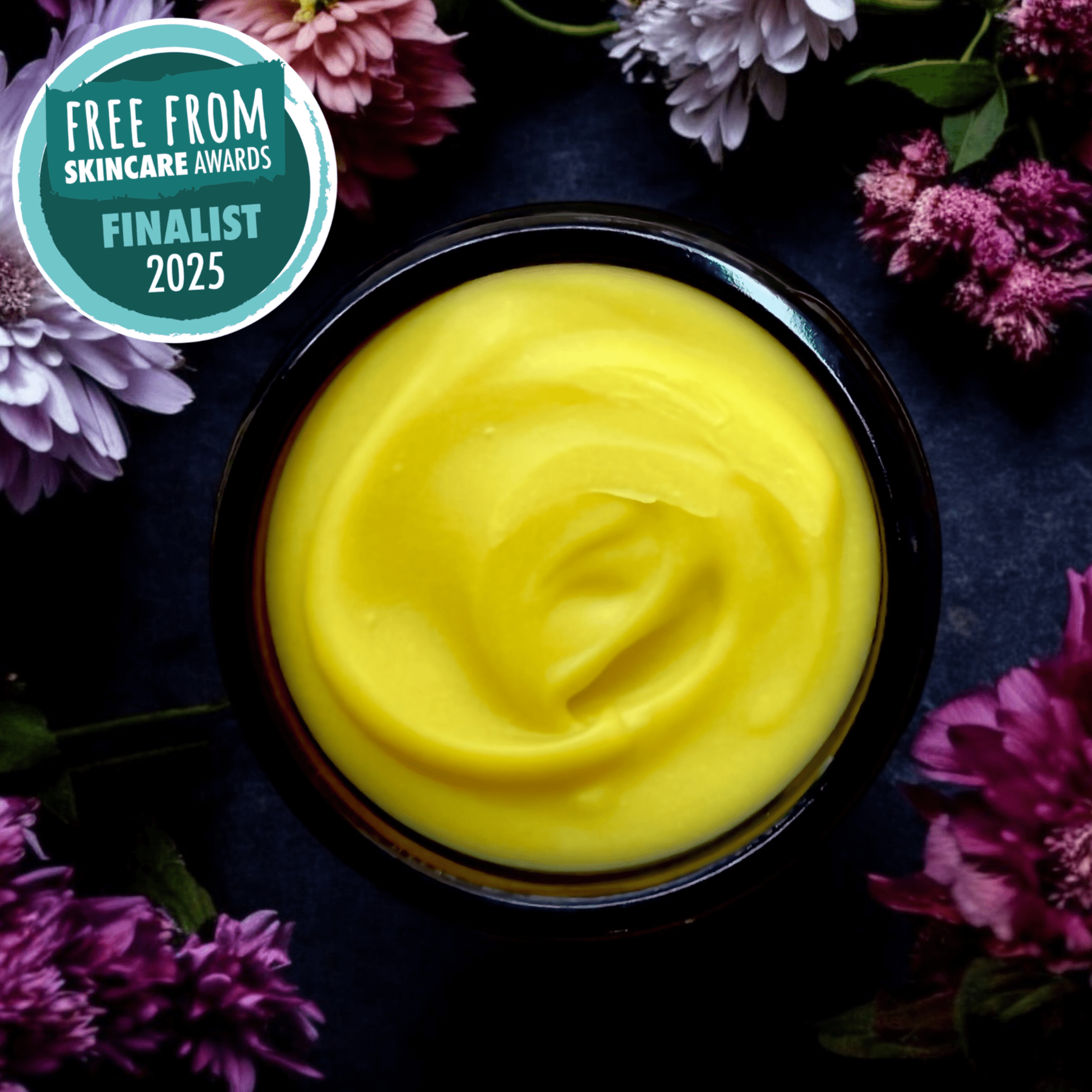 Yellow cream in a black container surrounded by purple flowers with 'Free From Skincare Awards Finalist 2025' badge.