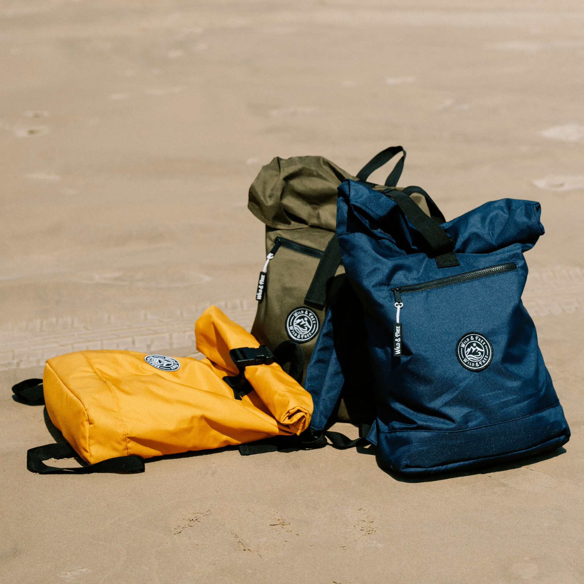 Three backpacks in yellow, green, and blue on a sandy surface.