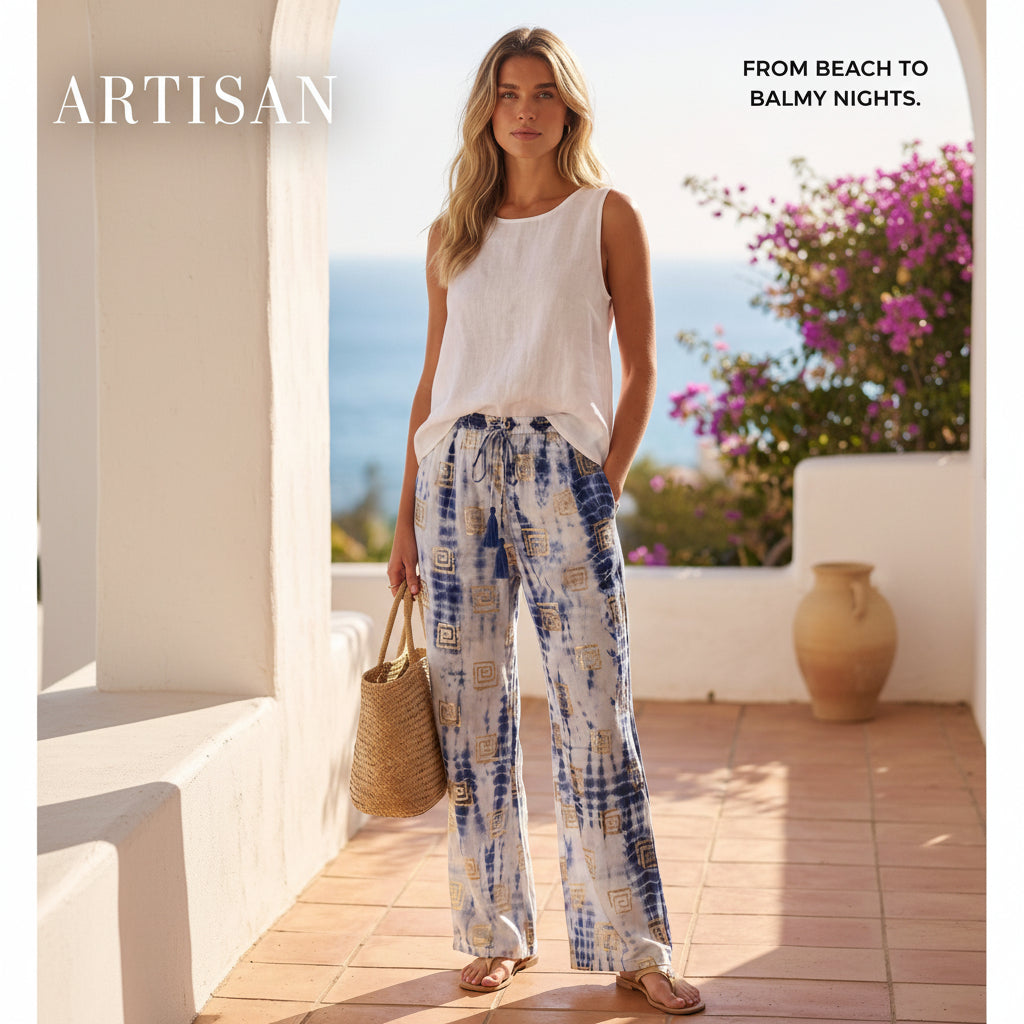 A woman stands on a sunlit terrace overlooking the ocean, wearing a white sleeveless top and loose white drawstring pants with blue and beige tie-dye patterns. She holds a woven straw bag and wears sandals.