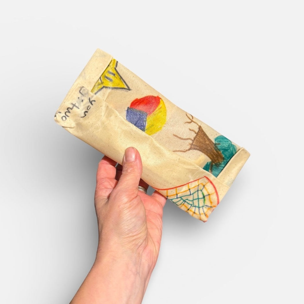 Hand holding a folded wax wrap with colourful drawings on a light grey background