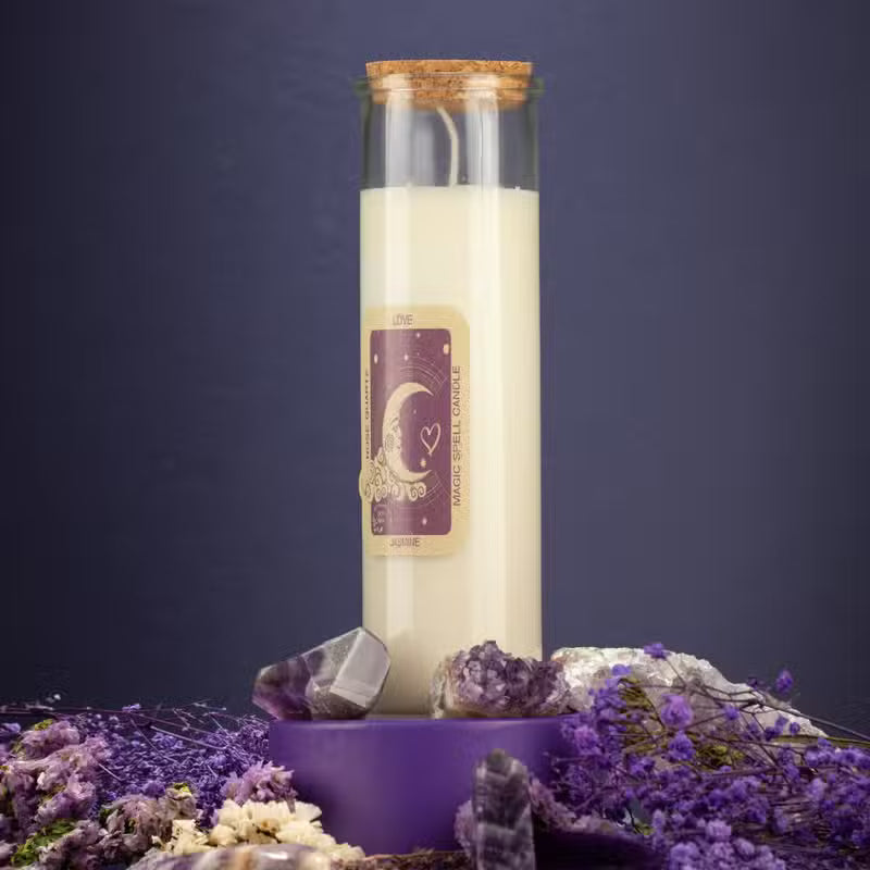 A tall glass jar candle with cream-colored wax and a cork stopper, featuring a purple and gold label with a crescent moon design. The candle is displayed on a purple pedestal surrounded by purple flowers, crystals, and dried botanicals against a dark purple background.