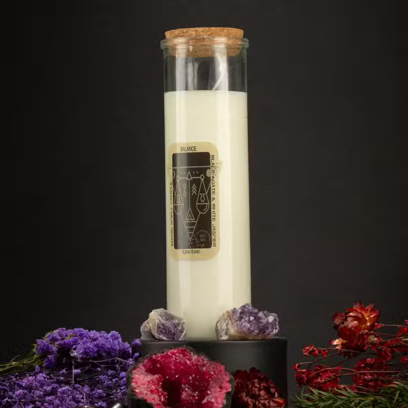 A white candle in a clear glass jar with a cork stopper, featuring a black and gold label that reads 'SILAGE' and 'Lunar Eclipse.' The jar is displayed on a black pedestal surrounded by purple, pink, and red crystals and dried flowers against a dark background.