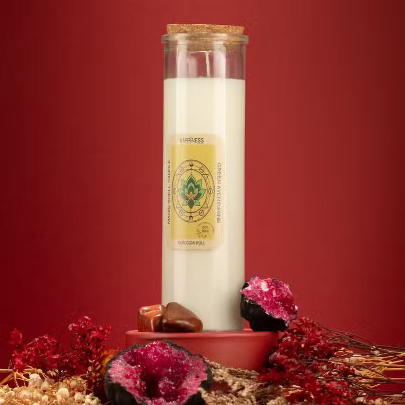 A white soy wax candle in a clear glass jar with a cork stopper and a cream-colored label reading 'HAPPINESS' and 'HAPPINESS BURNING SMOKE FRAGRANCE'. The candle is styled on a terracotta base with dried red flowers, brown stones, and geode props against a solid dark red background.
