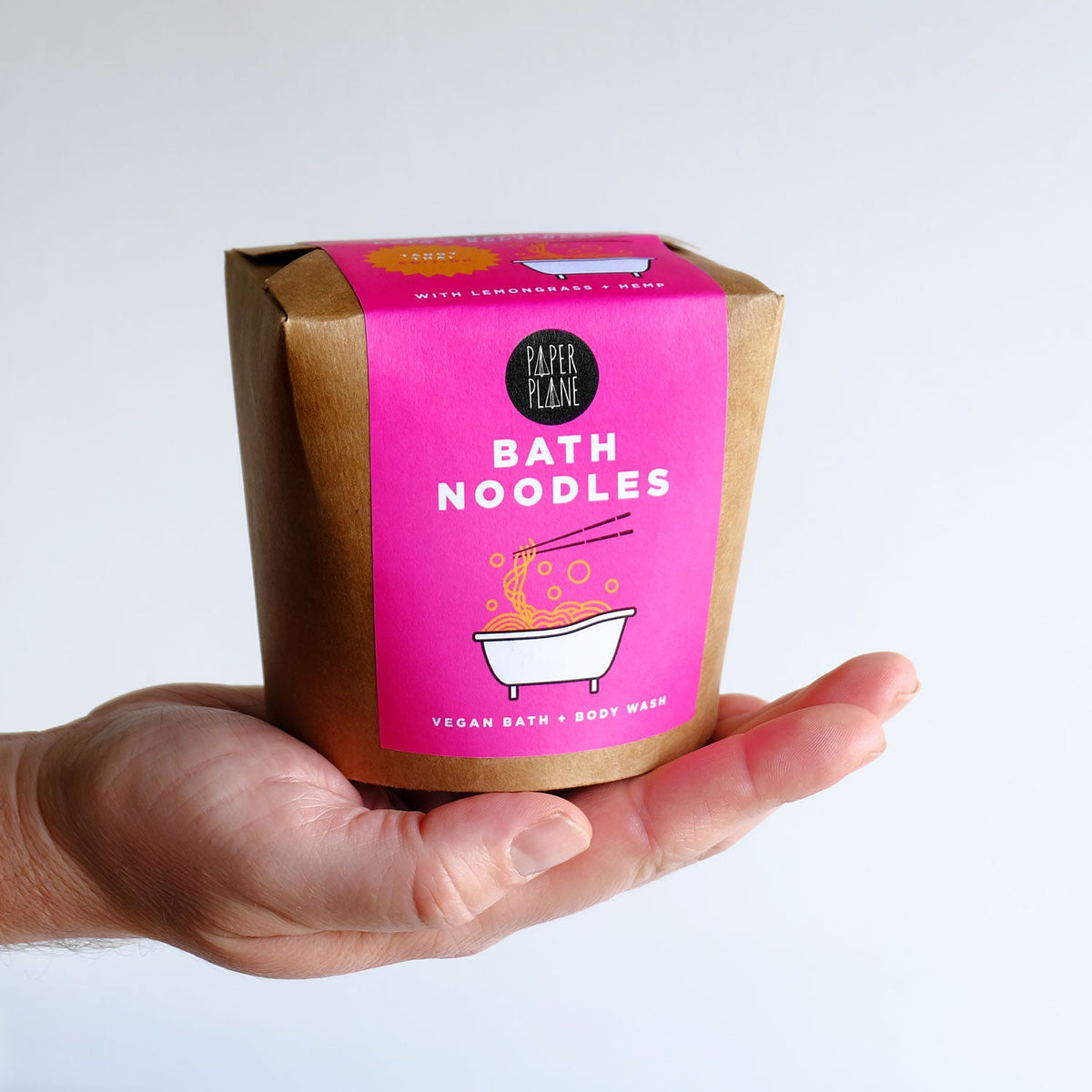 Hand holding a package of Paper Plane Bath Noodles with pink label and brown box.