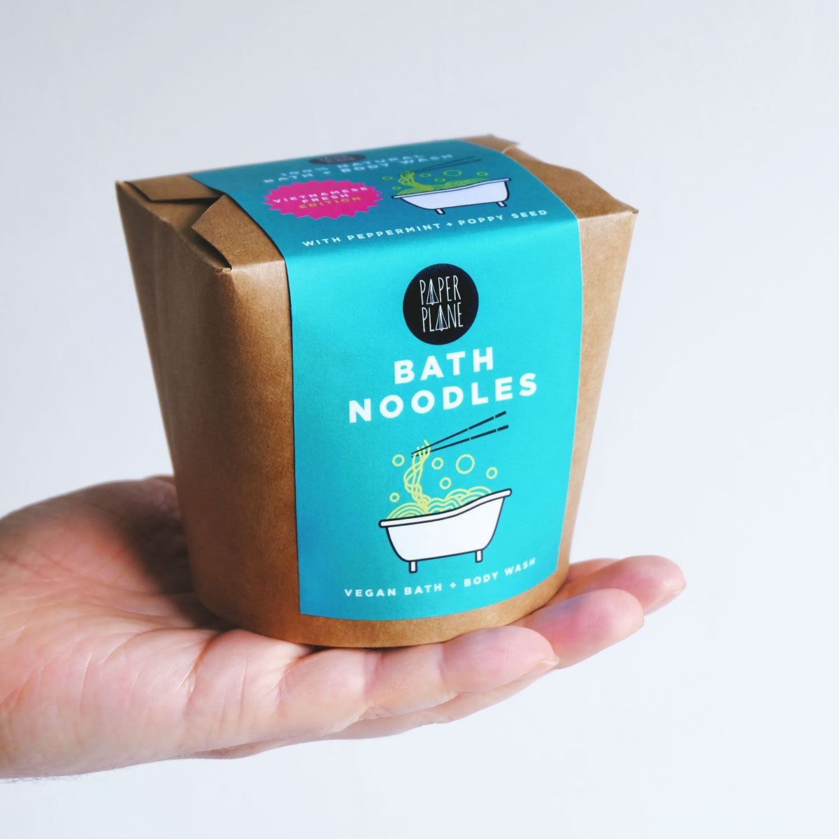 Hand holding a package of Paper Plane Bath Noodles with a blue label.