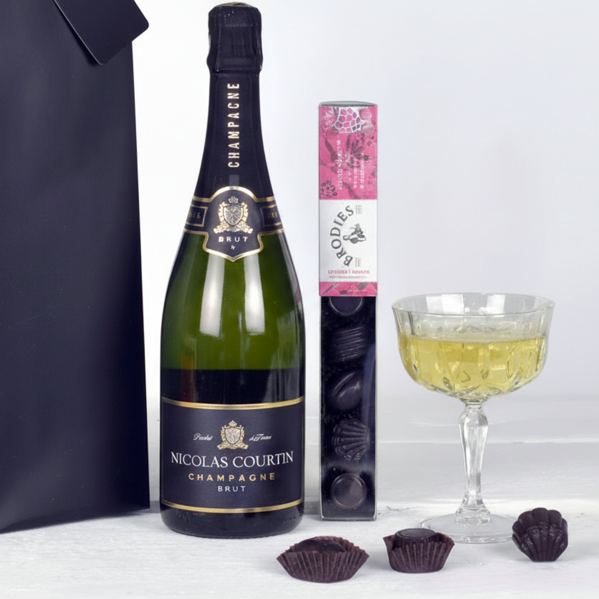 Nicolas Courtin champagne bottle with a box of Godiva chocolates and a glass of champagne on a white surface.
