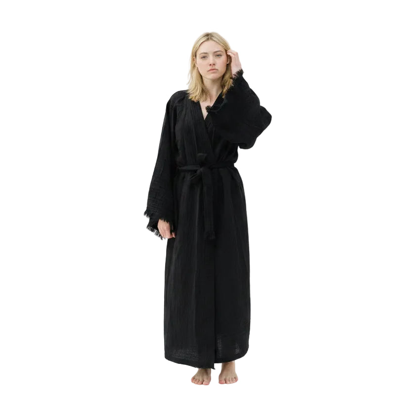 A woman modelling a long black organic cotton robe with wide sleeves and a tie belt, standing barefoot against a plain white studio background.