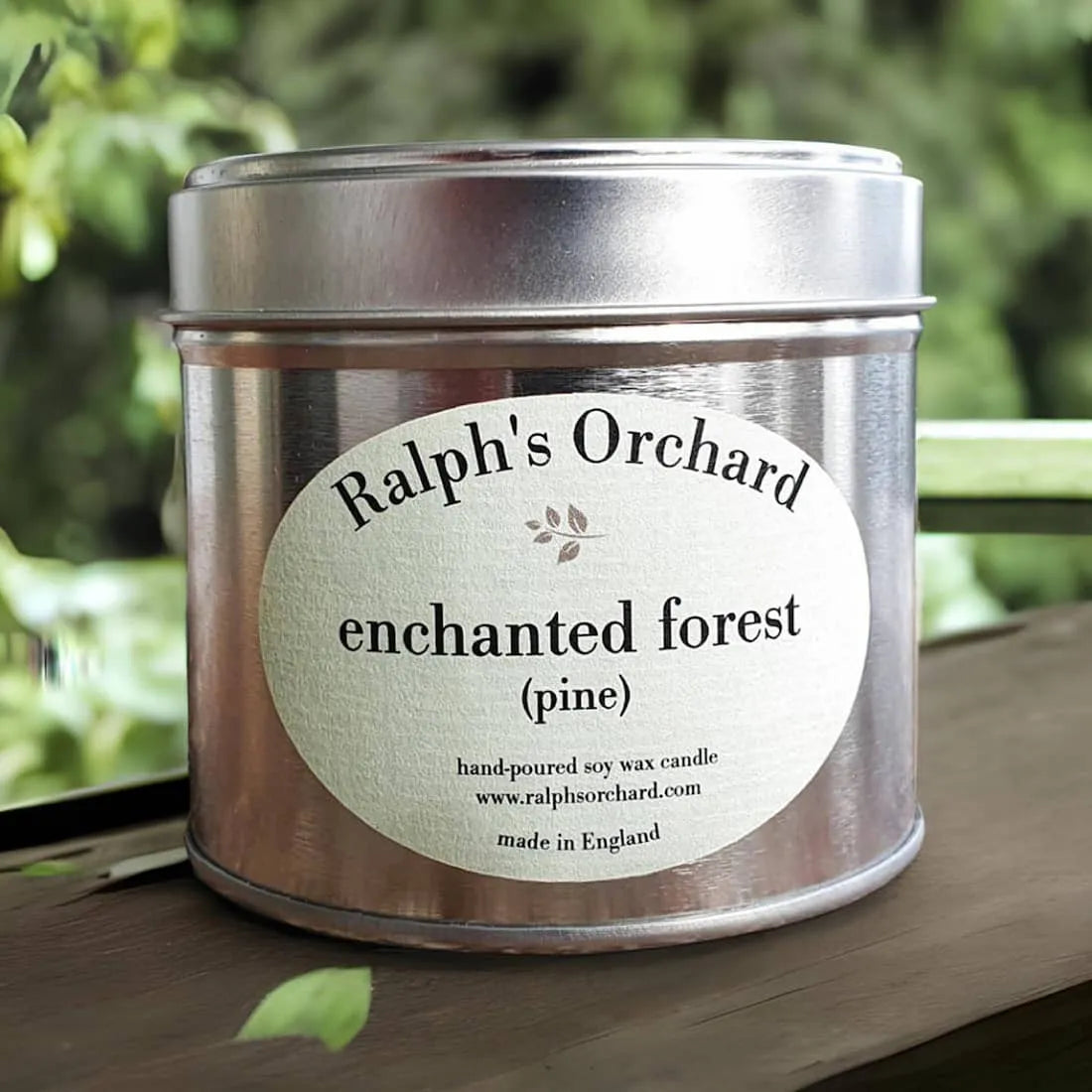 Candle tin labeled 'Ralph's Orchard Enchanted Forest Pine' on a wooden surface with a blurred natural background.