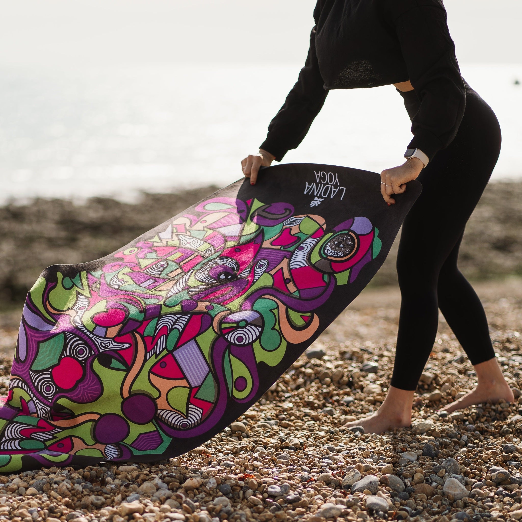 Person holding a colourful yoga mat with a pattern on a pebbly beach.