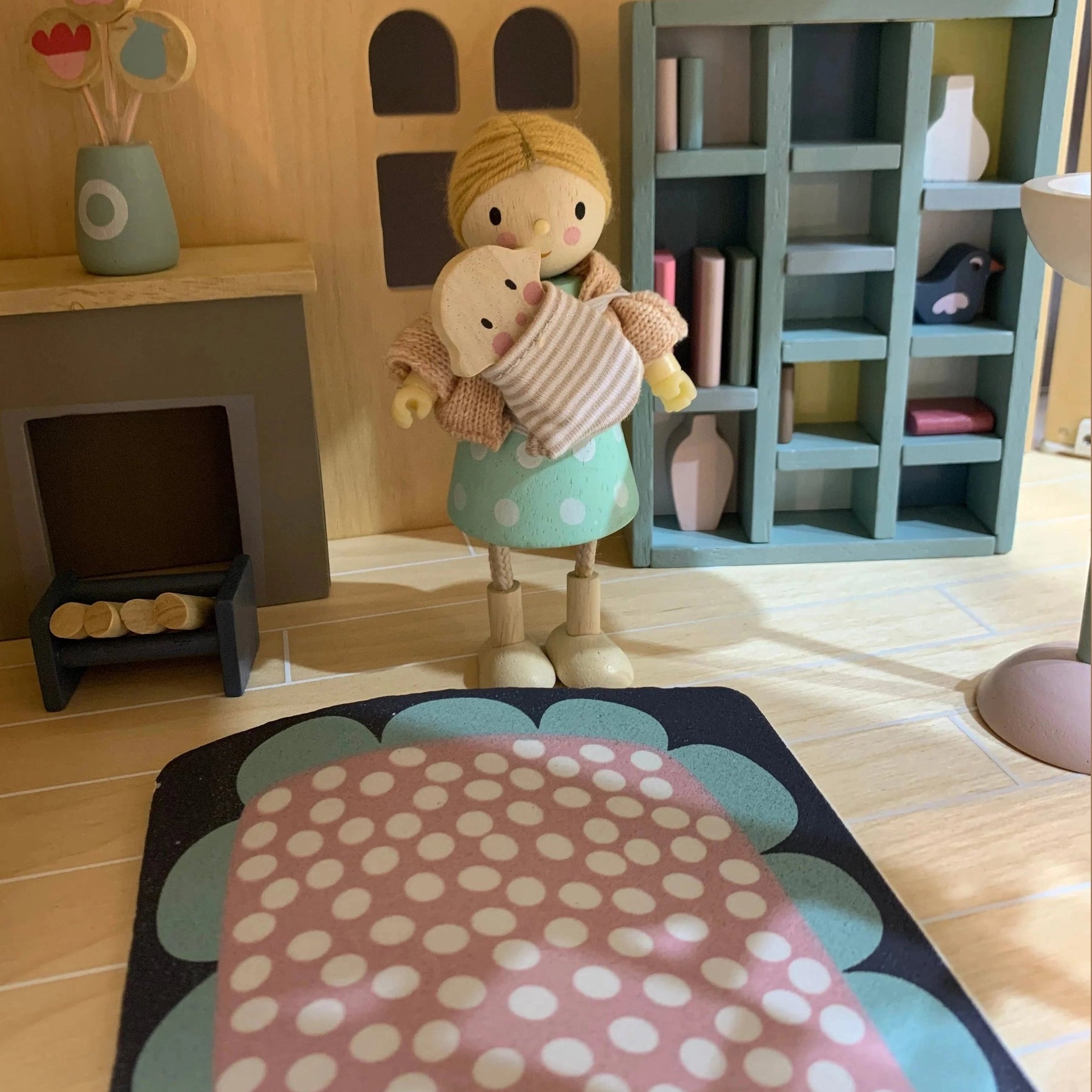 Wooden doll holding a baby doll in a doll's house living room with a bookshelf and fireplace.