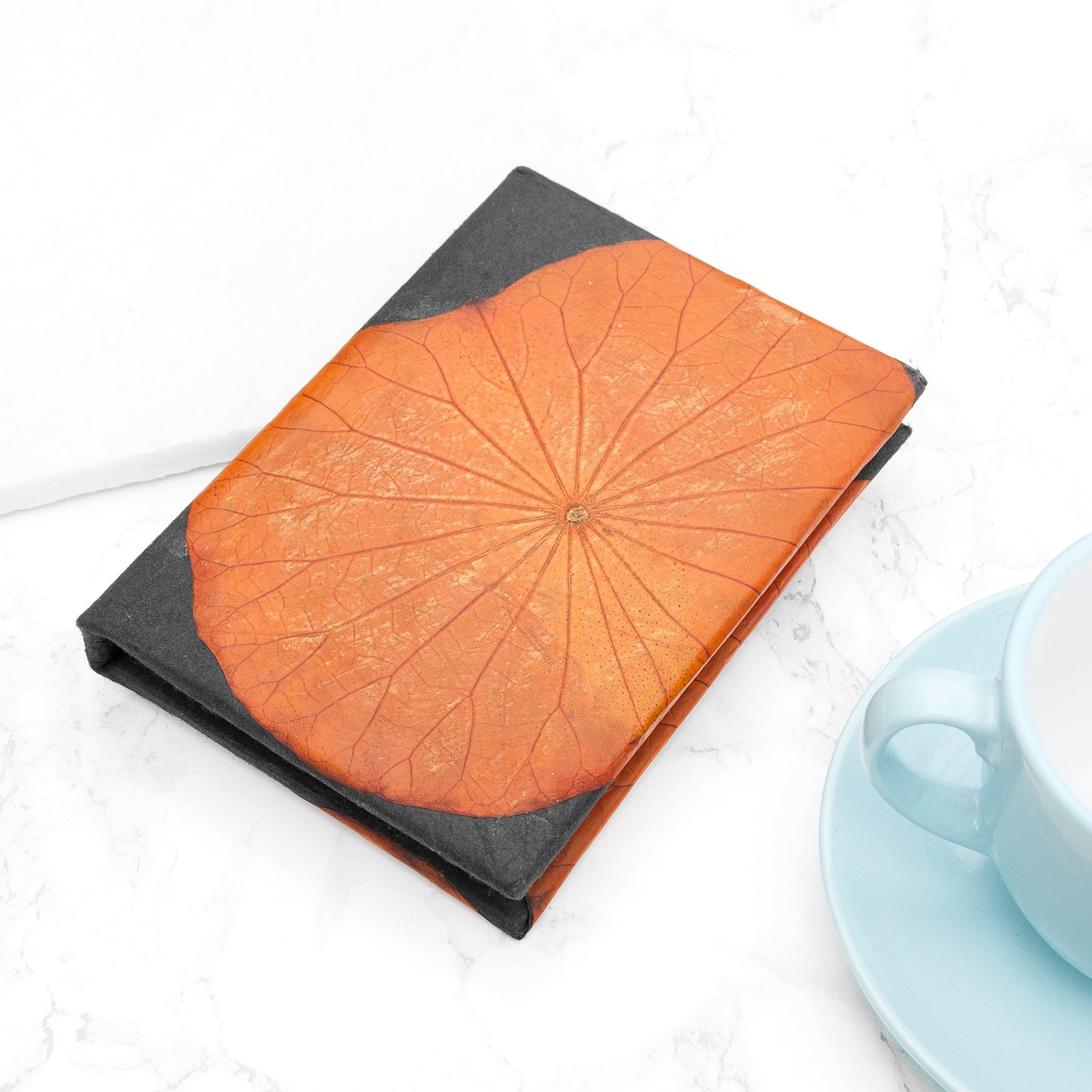 Notebook with orange leaf pattern cover on a white surface with a blue cup and saucer.