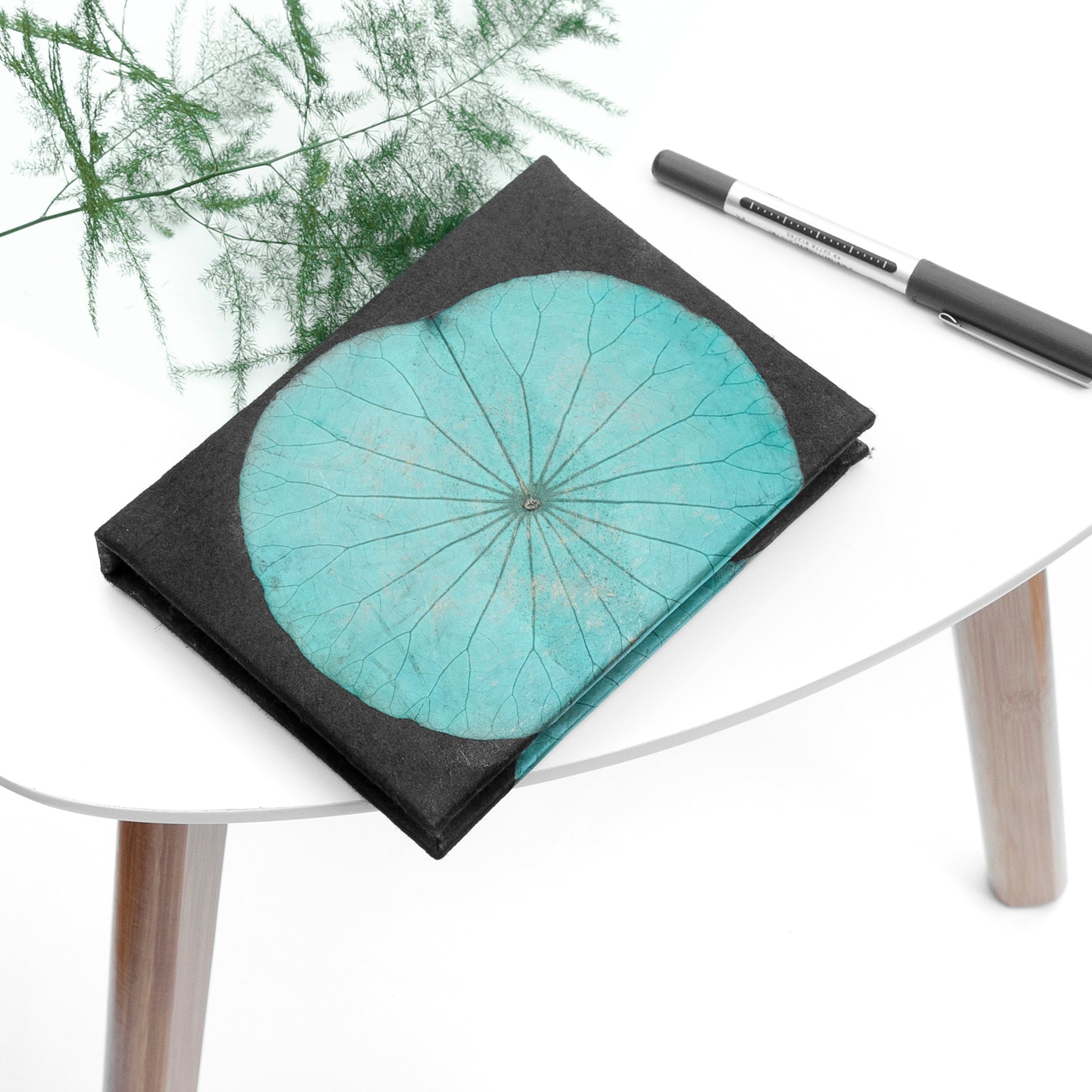 Teal floral design on a black notebook with a pen next to it on a white surface.