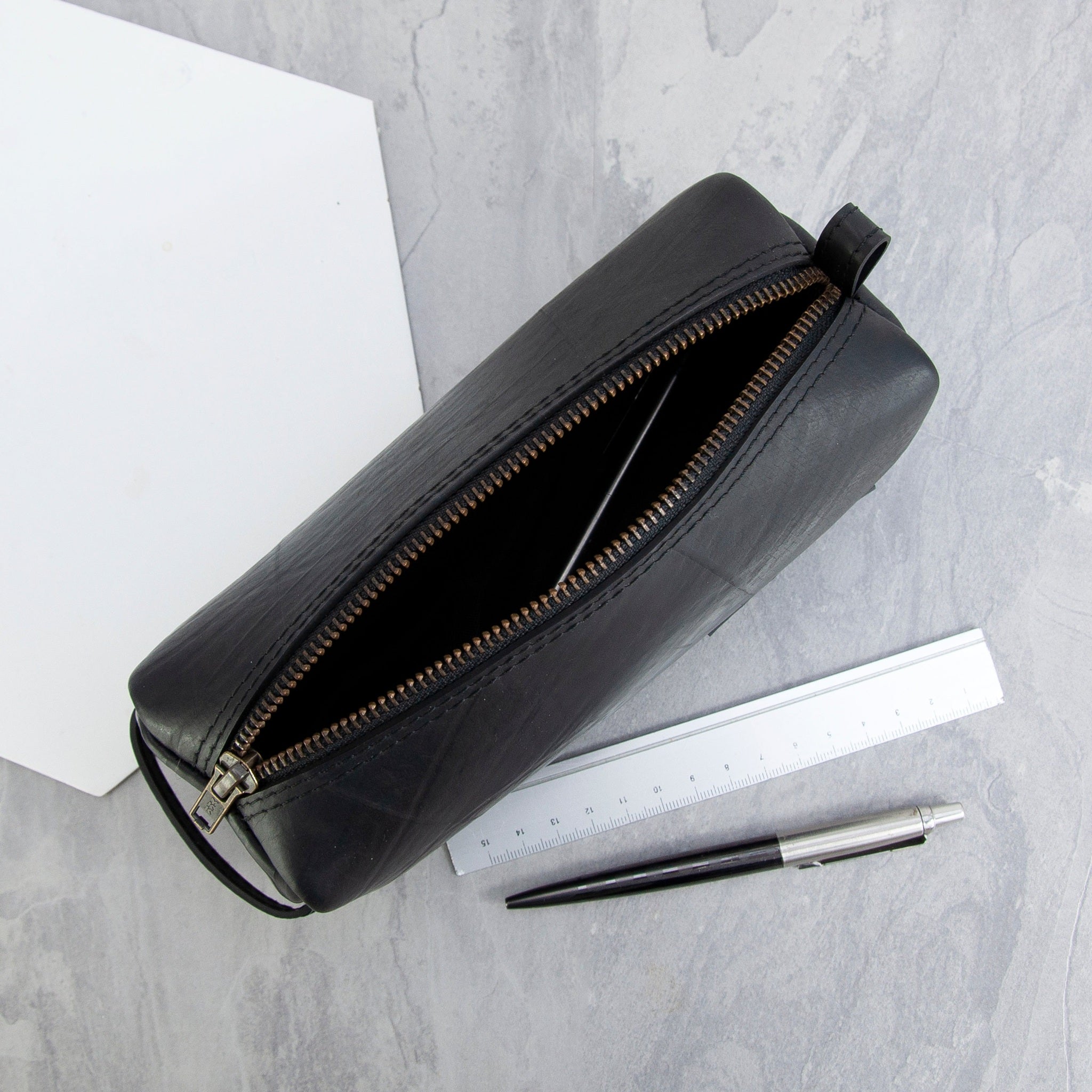 Black recycled rubber pencil case with a gold zipper on a gray surface with a pen and paper.