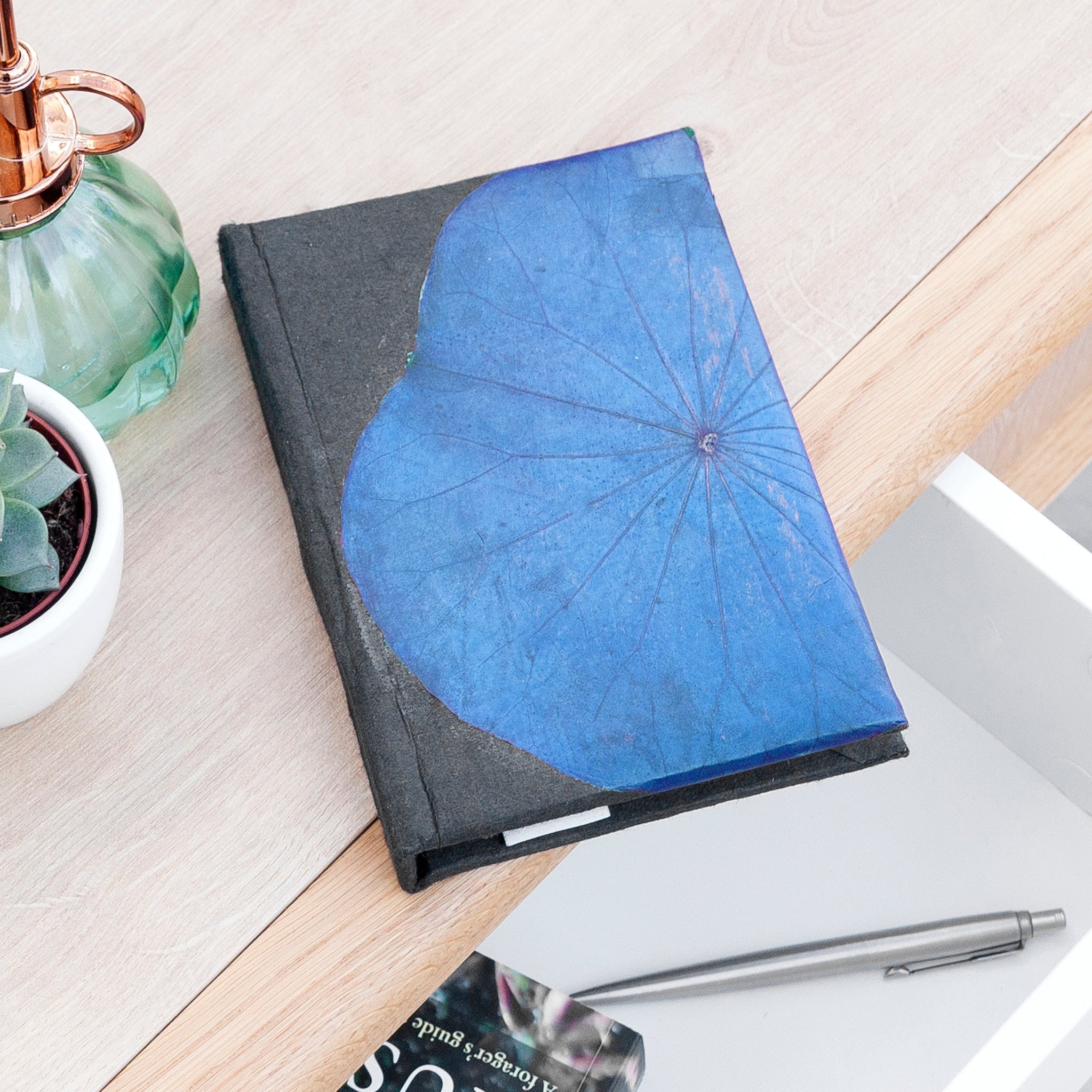 Notebook with blue flower design on cover, placed on a wooden surface with a pen and plant in the background.