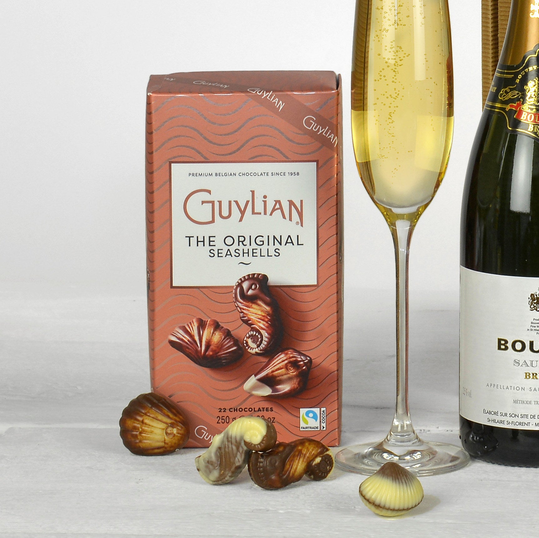 Box of Guylian chocolate seashells with a glass of fizz and a bottle of fizz on a light background
