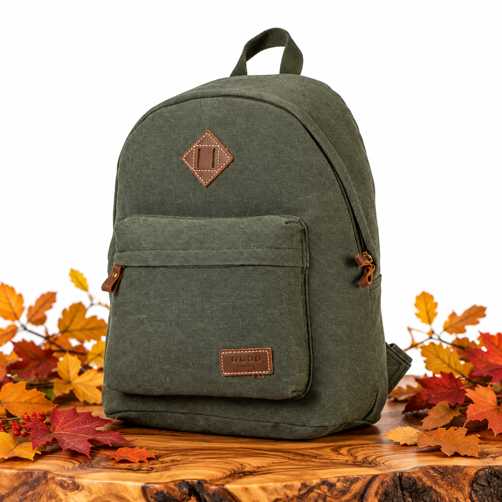 An olive green canvas backpack with brown leather accents sits on a wooden surface surrounded by colorful autumn leaves against a white background.