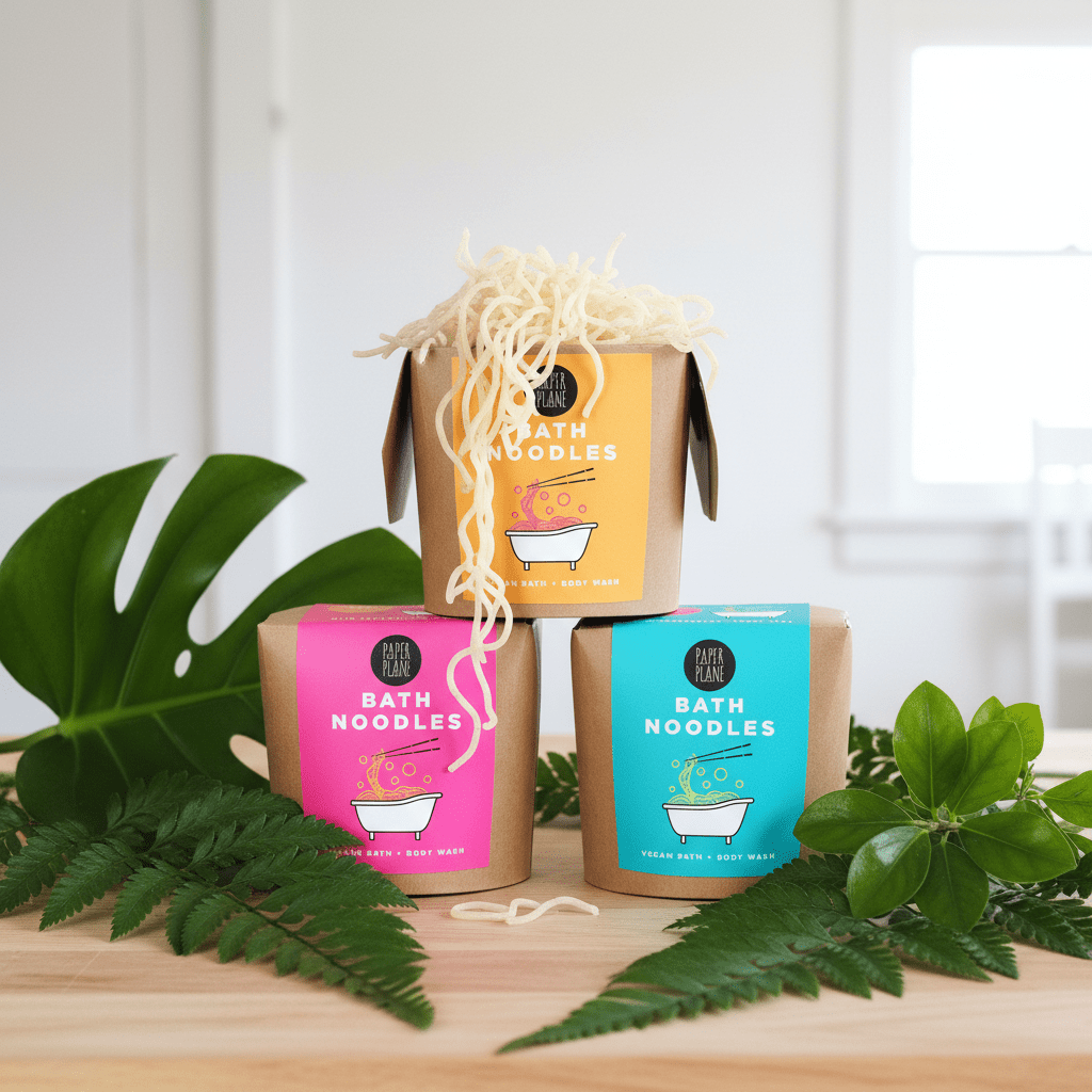 Three packages of Bath Noodles with colorful labels on a wooden surface with green leaves.