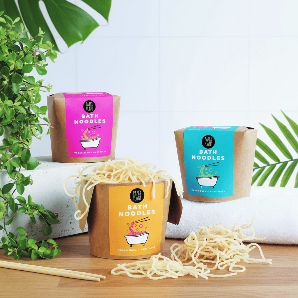 Three packages of bath noodles with visible noodles on a wooden surface, surrounded by green leaves.