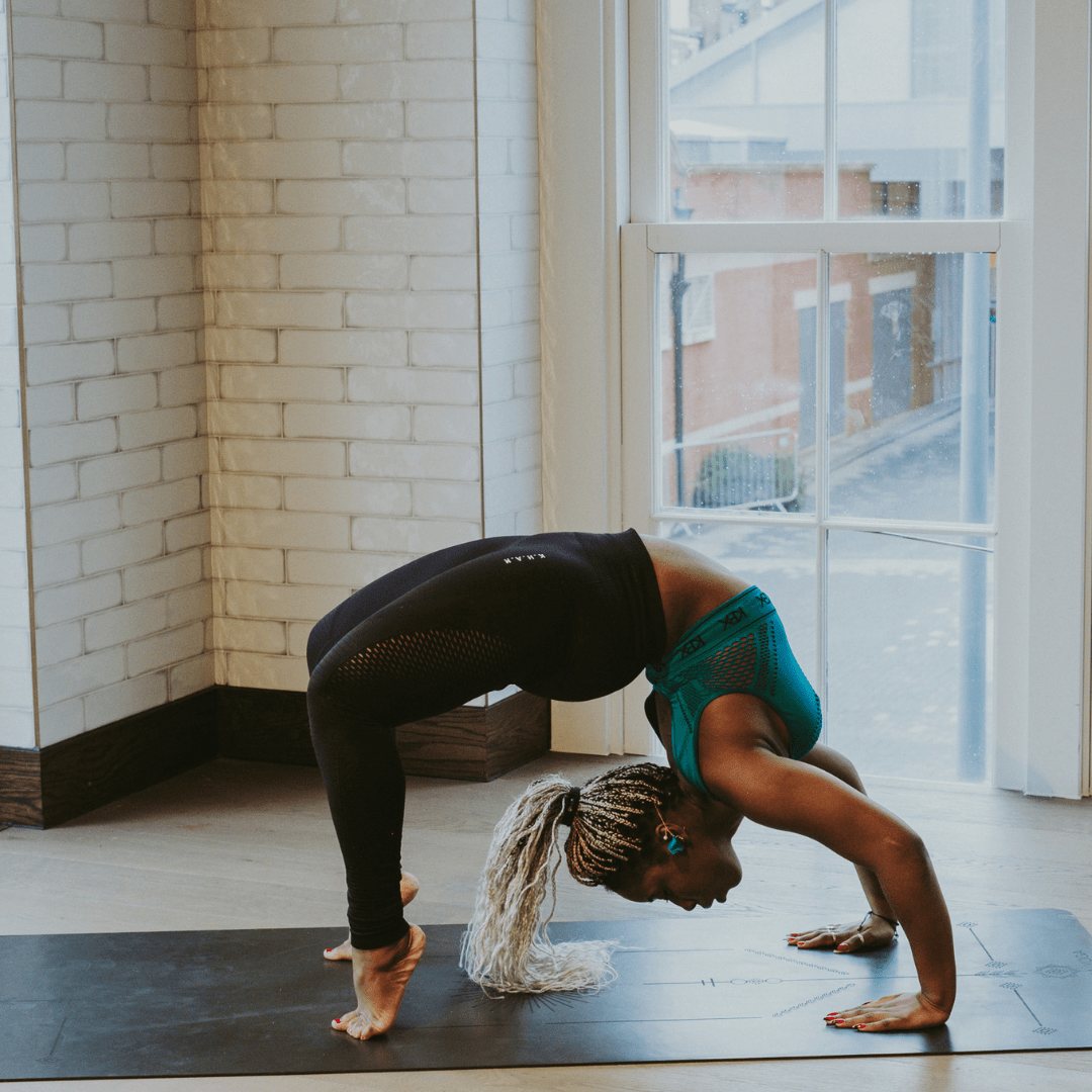 A person performing a yoga pose on a black yoga mat in a room with large windows and a brick wall.