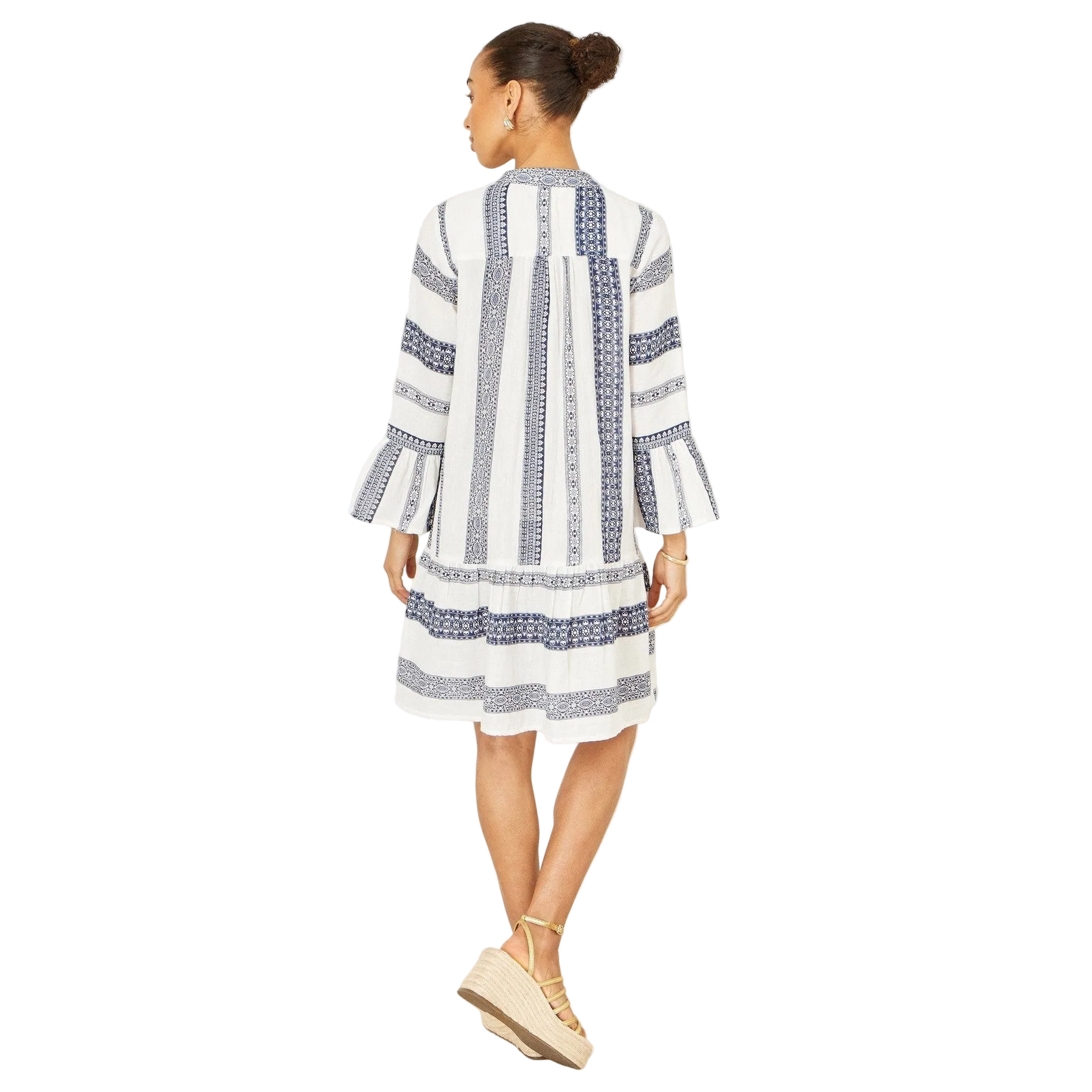 A woman shown from the back wearing a white long-sleeve dress with vertical navy blue patterned stripes and beige lace-up wedge sandals, set against a plain white background.
