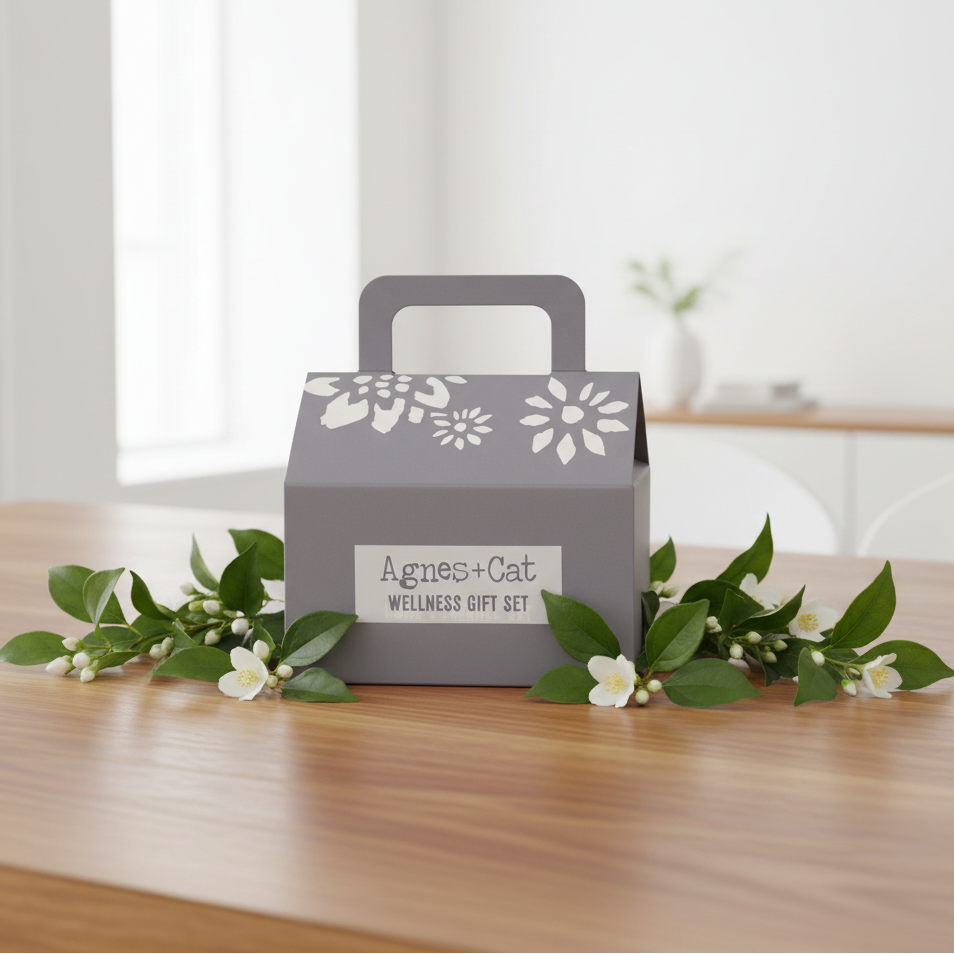 A grey gift box with a handle, decorated with white floral patterns and a label reading 'Agnes+Cat WELLNESS GIFT SET,' sits on a wooden table surrounded by green leaves and small white flowers in a softly lit indoor setting.