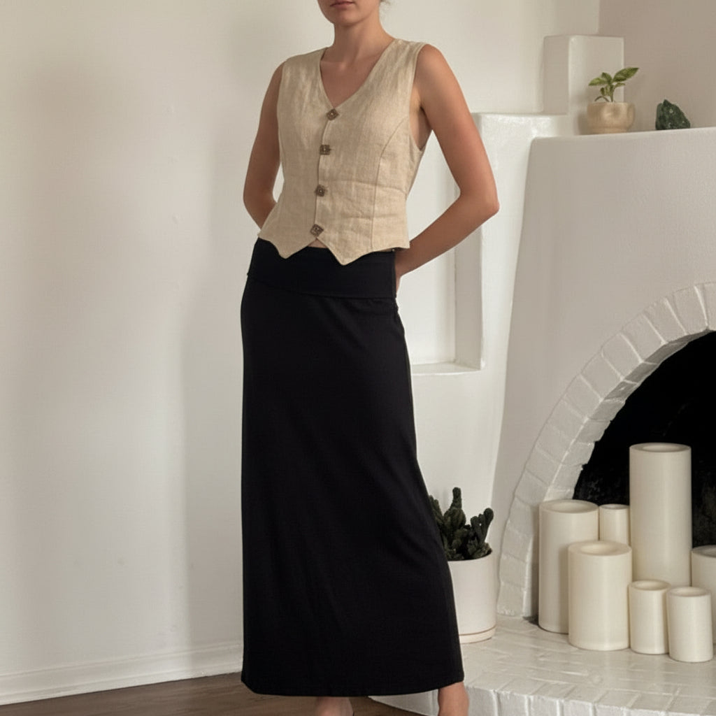 Woman wearing a beige vest and black skirt standing in a room with a fireplace.