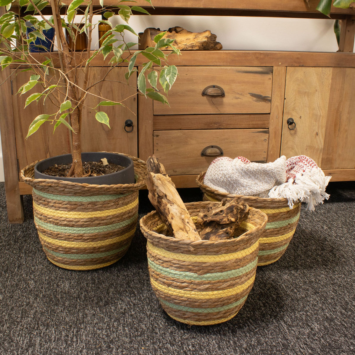 Three striped baskets holding a potted plant, folded textiles and logs beside a wooden sideboard