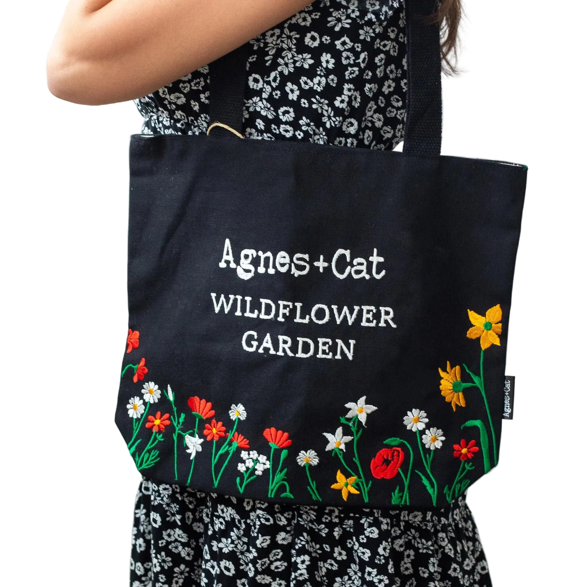 Person holding a black tote bag with floral designs and 'Agnes + Cat Wildflower Garden' text.
