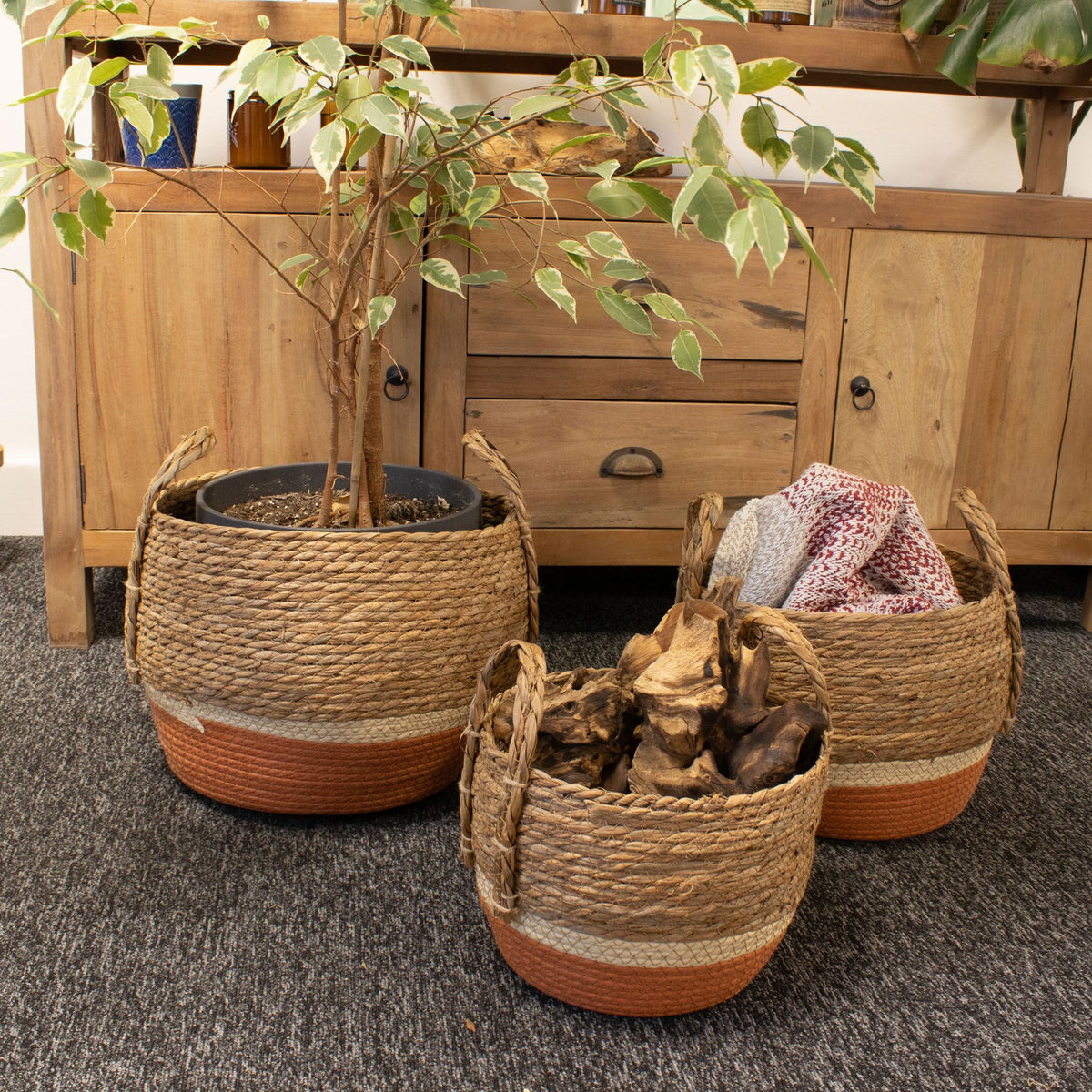 Three orange-based baskets holding a potted plant, folded textiles and logs beside a wooden sideboard