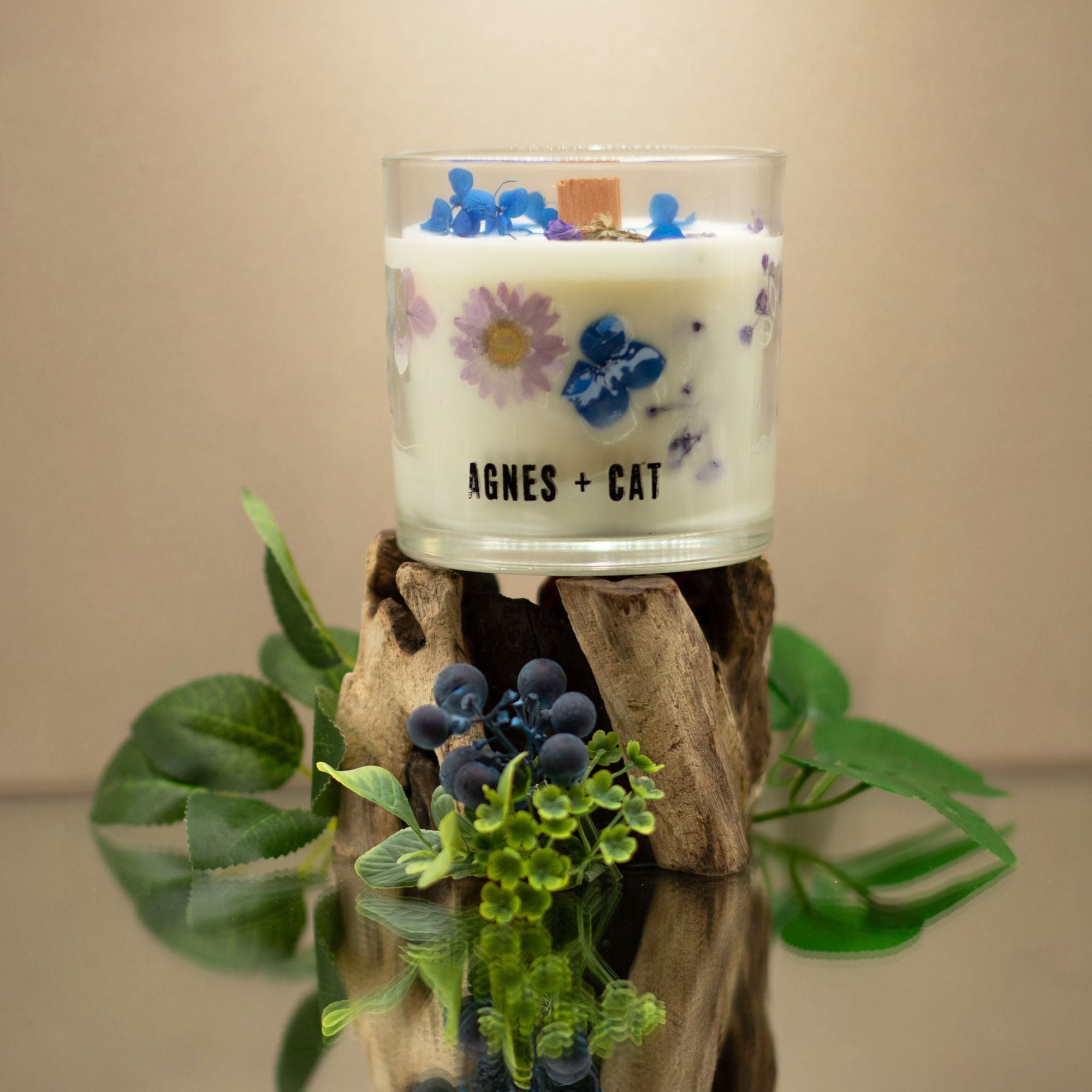 Candle with floral design and 'AGNES + CAT' branding on a wooden stand with plants.