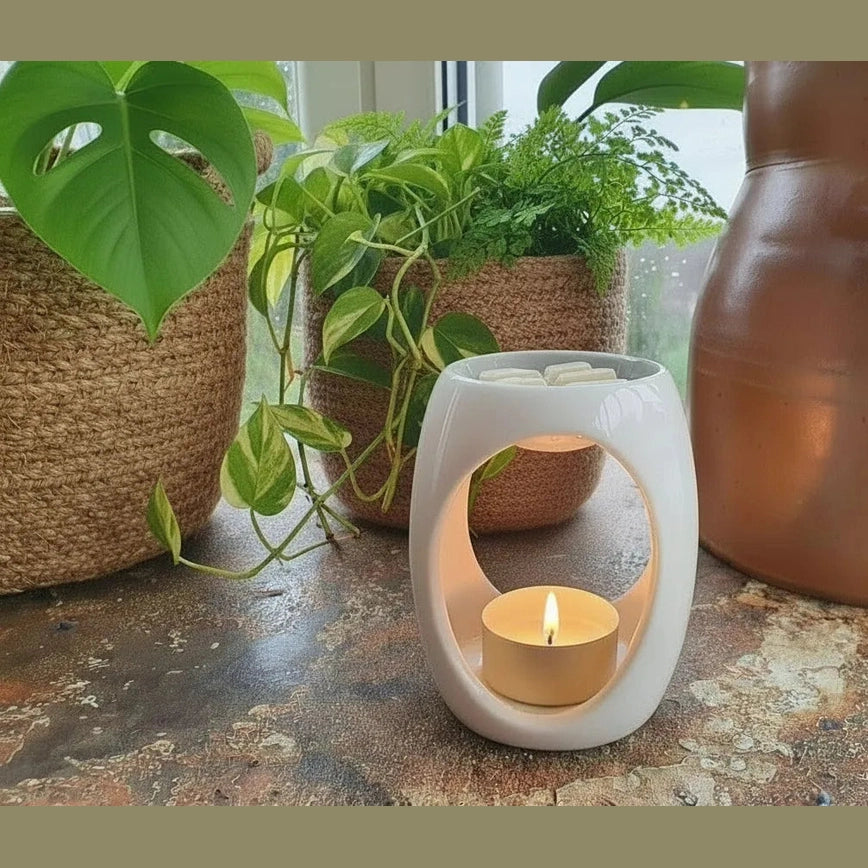 White ceramic oil burner with a lit candle on a stone surface with plants in the background