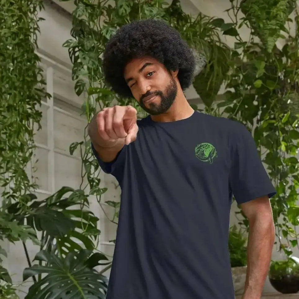 A man wearing a navy blue t-shirt with a green logo, standing in front of greenery.