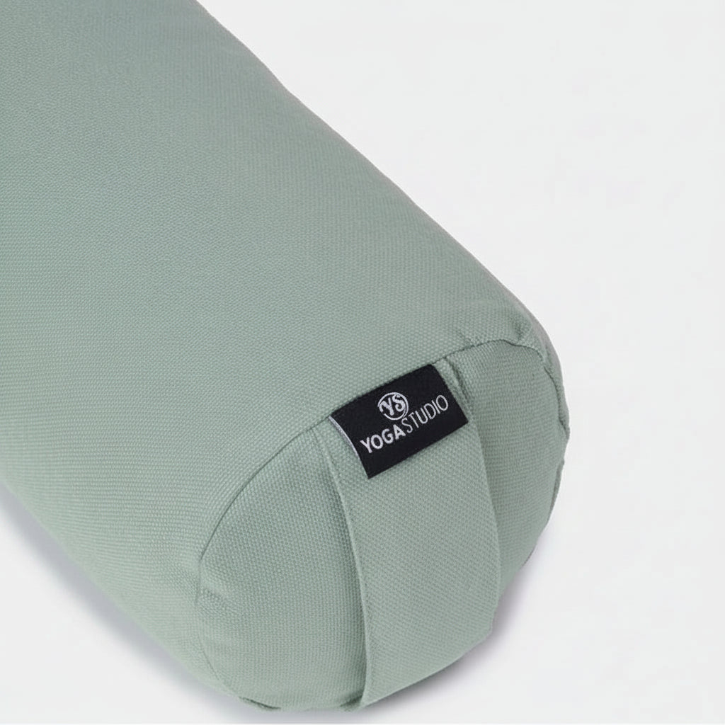 A sage green fabric yoga bolster pillow with a black fabric brand label reading 'YOGASTUDIO' on a light gray background.