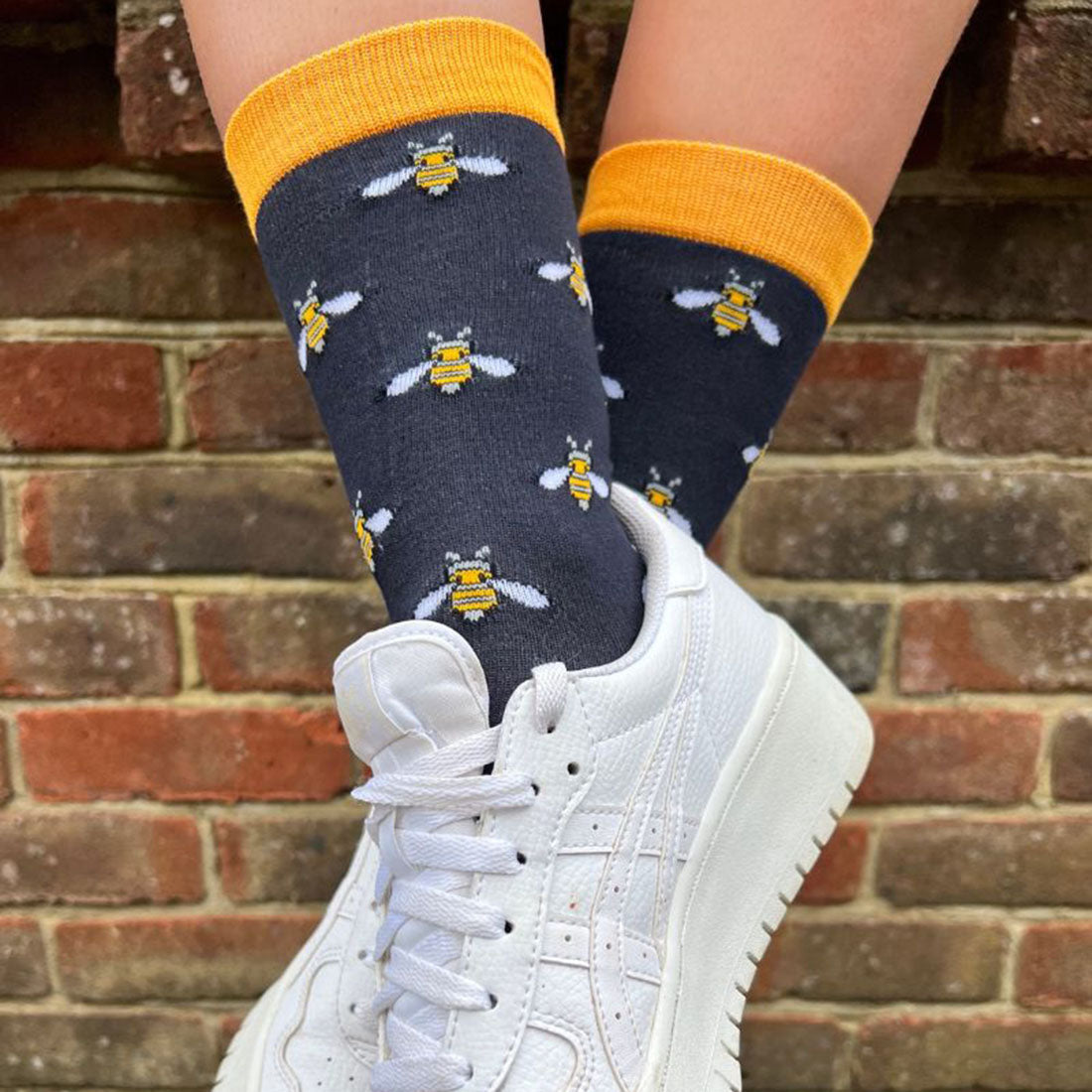 Person wearing navy socks with bee patterns and white sneakers against a brick wall.