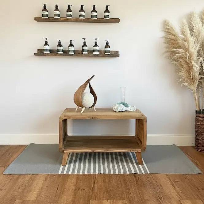 A styled interior corner with a wooden console table, two floating wall shelves holding small bottles, a decorative coconut lamp, a striped rug, and pampas grass against a white wall.