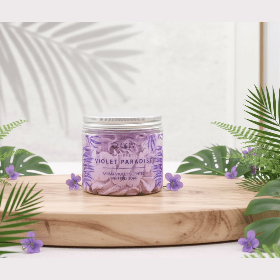 A jar of whipped cream soap labelled 'Violet Paradise' on a wooden board with greenery