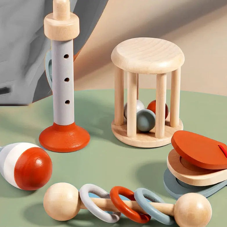 A set of wooden baby sensory toys arranged on a soft green surface, including a ring rattle, a small wooden stool with hanging balls, a vertical peg toy, and a clacker.
