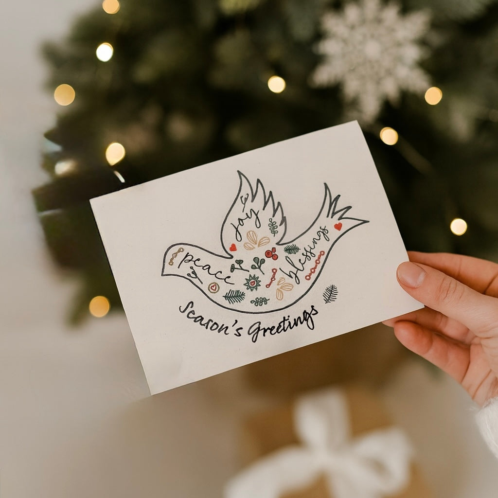 Hand holding a Christmas card with a dove design and 'Season's Greetings' text, against a festive background.