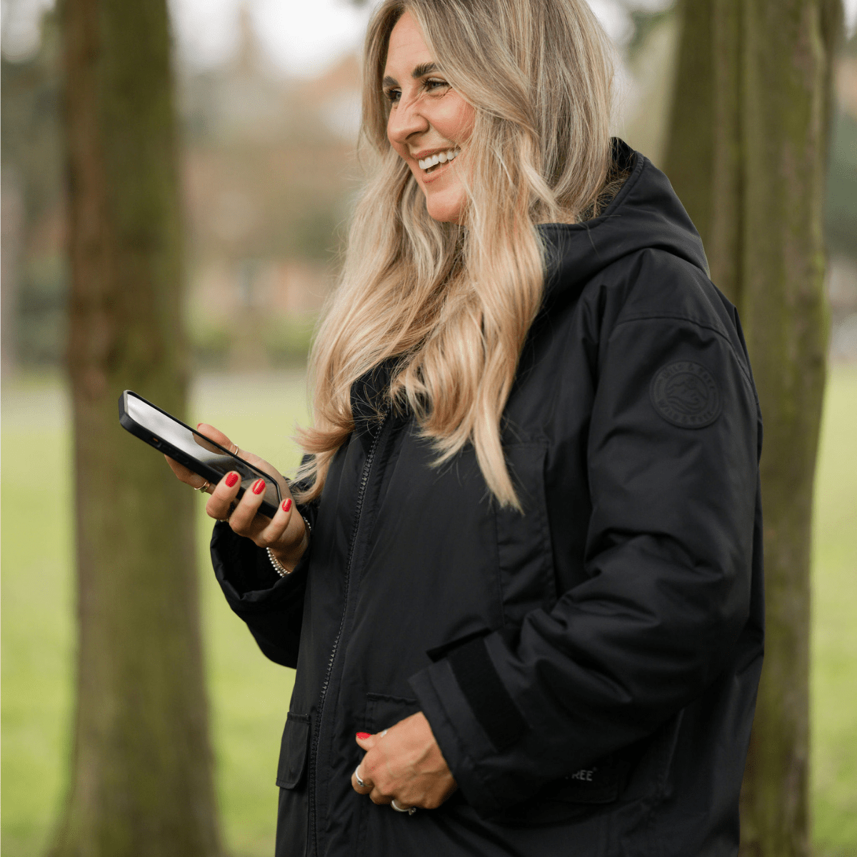 Woman in a black coat holding a phone outdoors with trees in the background