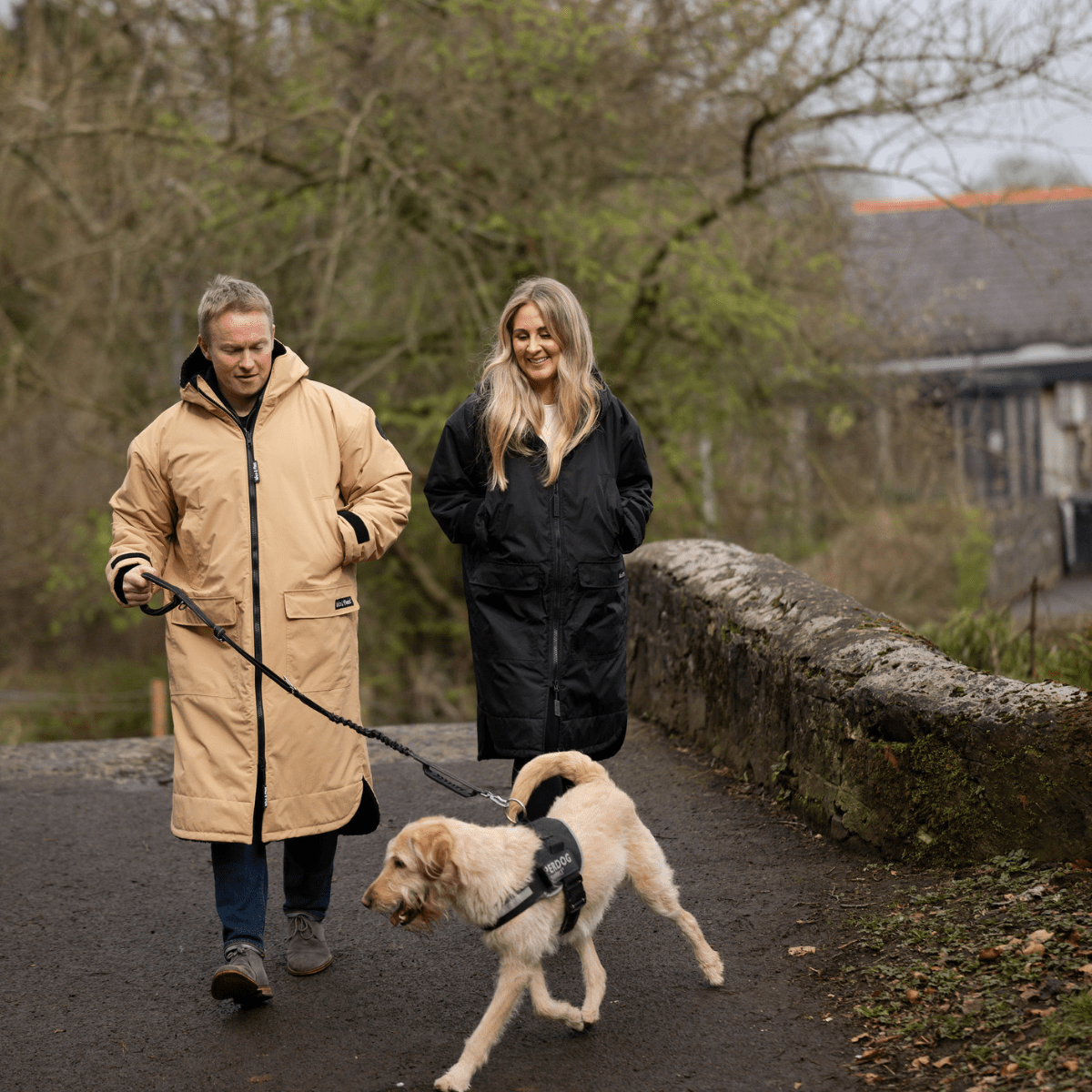 Two people walking a dog on a path with trees and a building in the background