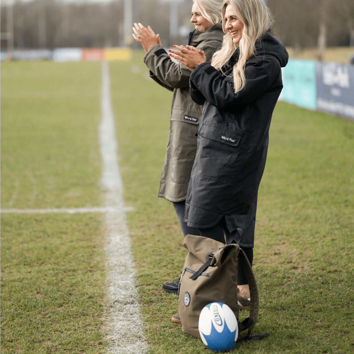 Two women standing on a grassy field with a sports ball and bag, clapping.