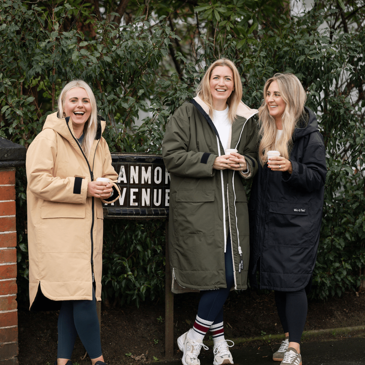 Three women in coats standing outdoors holding drinks, with a sign in the background.