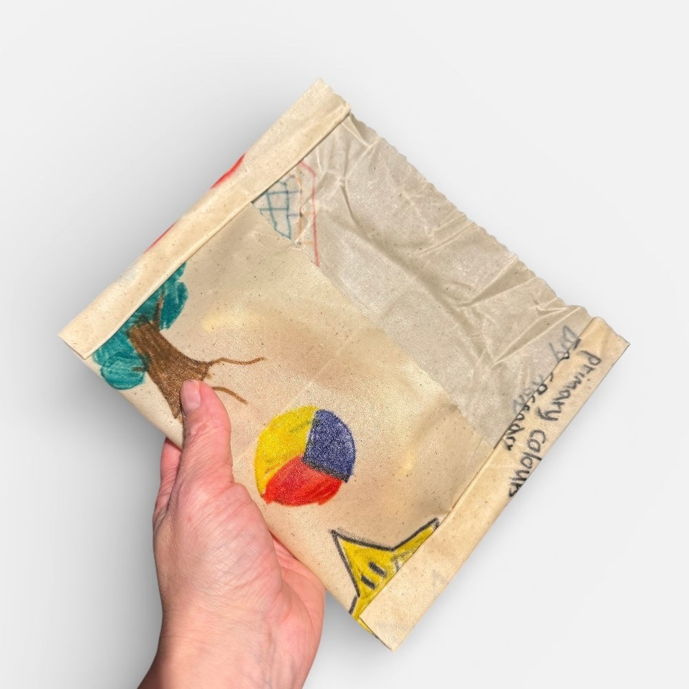 Hand holding a small wax wrap with colourful designs on a light grey background